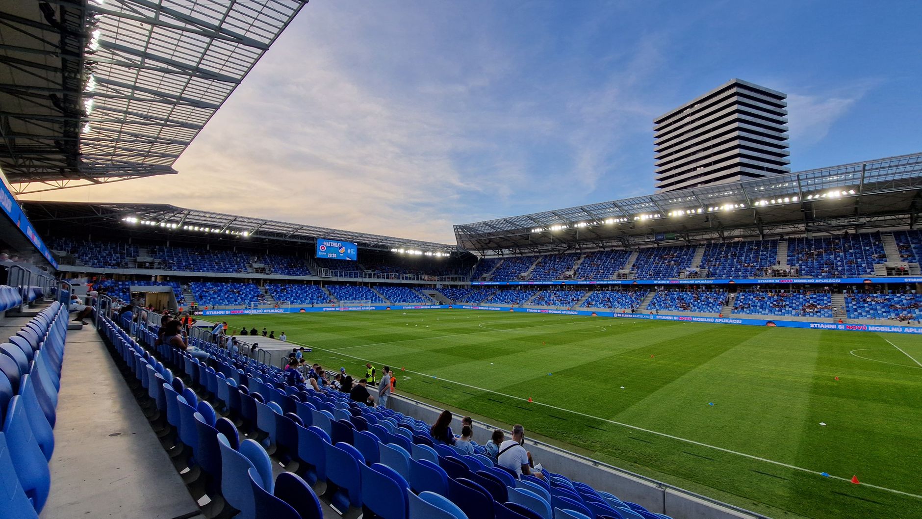 A panoramic view of a modern football stadium in Bratislava, Slovakia during dusk with blue seating and a grassy field.