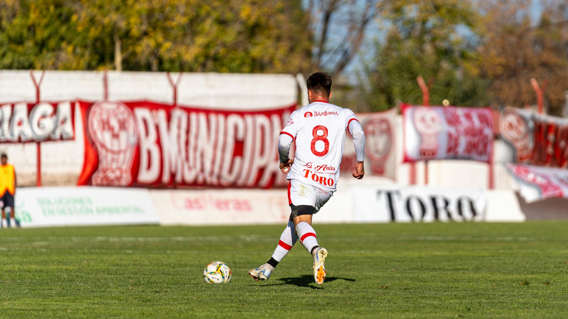 A soccer player in a white uniform dribbles the ball during a sunny outdoor match.