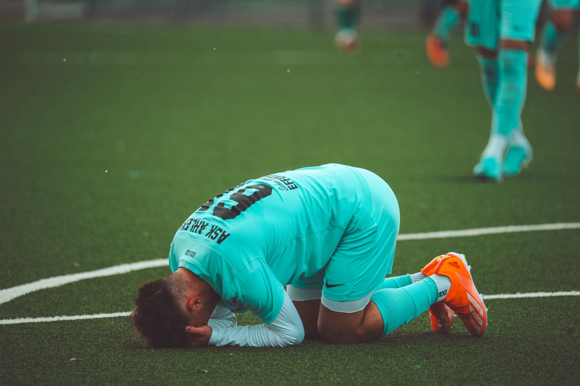 A soccer player kneels in despair on a green artificial turf sports field.