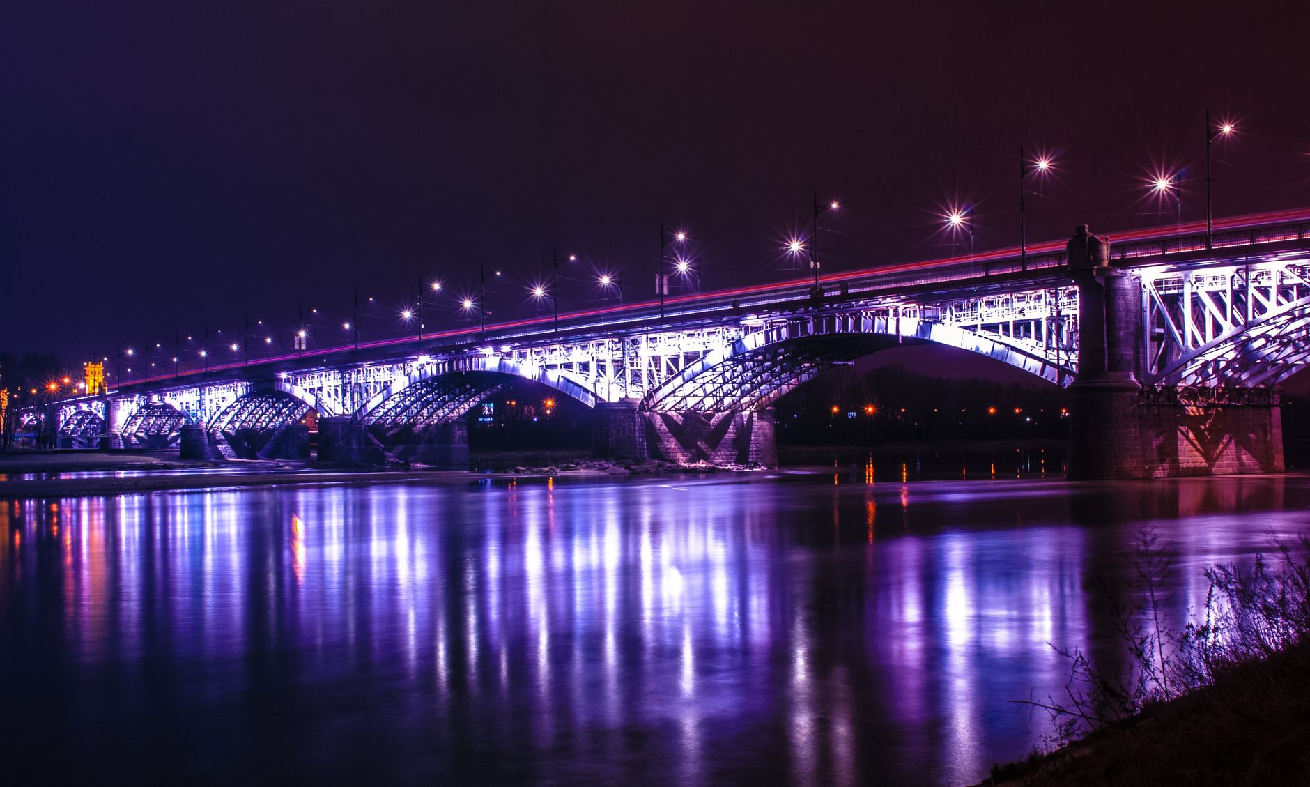 A stunning night view of an illuminated bridge reflecting on the Wisła River in Warsaw, Poland.