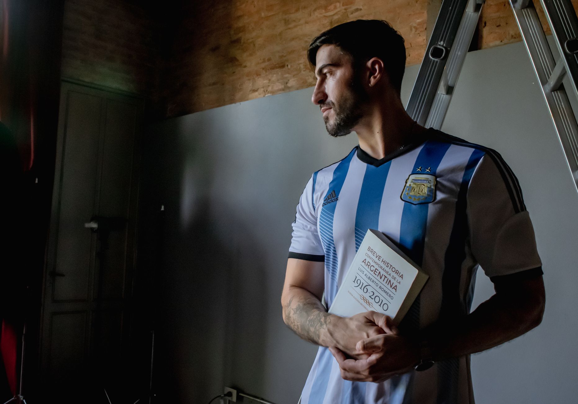 A man in an Argentina soccer jersey holds a history book indoors. Brick wall background.