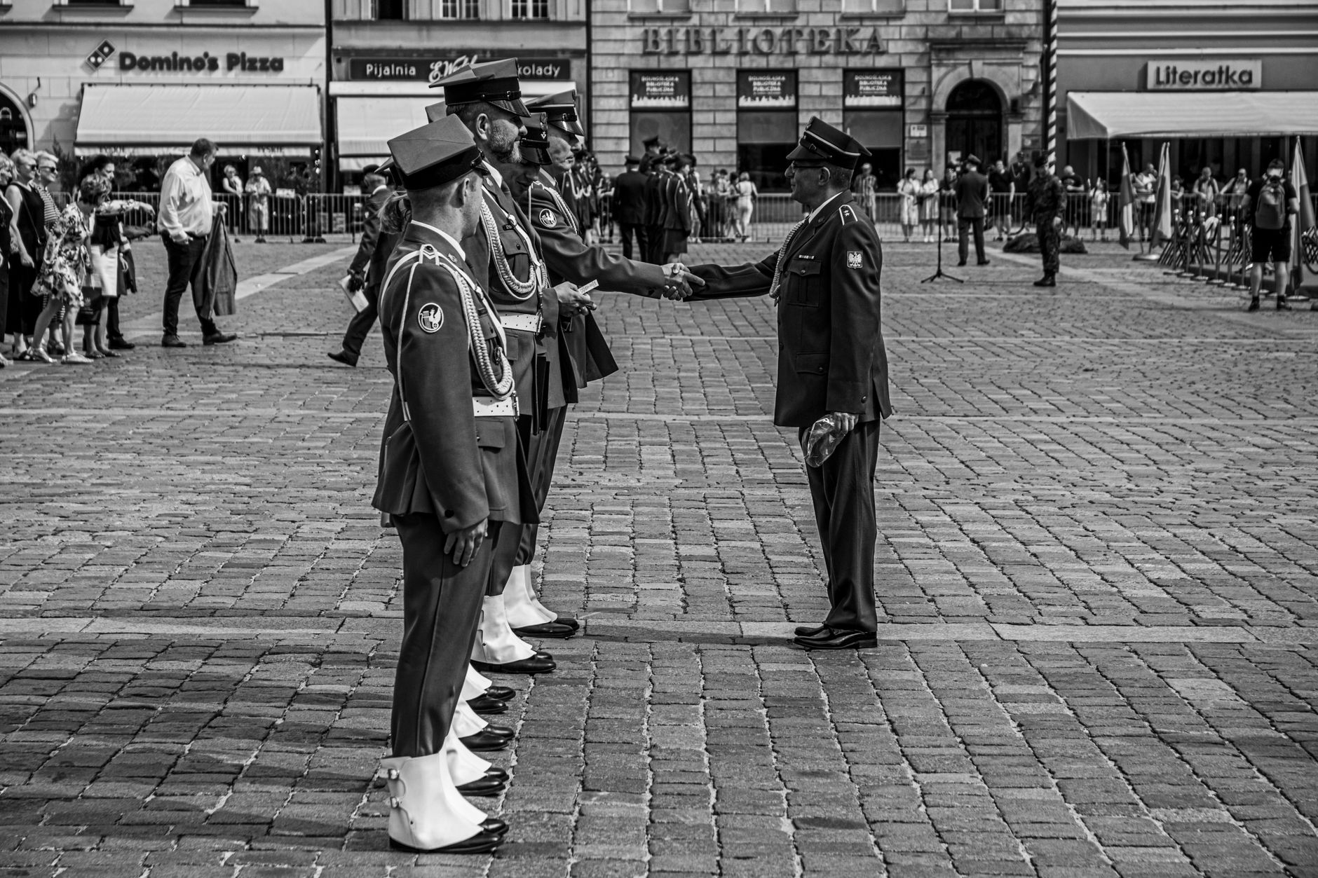 Formal handshake between uniformed officers in a ceremonial setting on a cobblestone square.