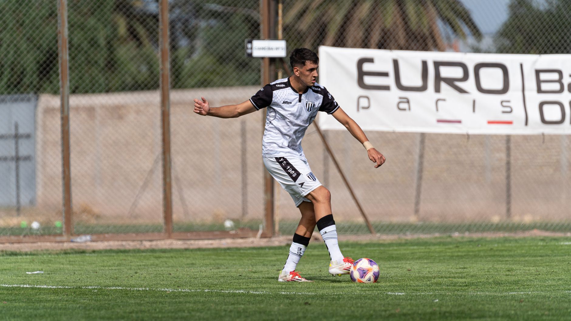 A soccer player in a white and black uniform dribbling a ball on a grass field during a daytime outdoor match.