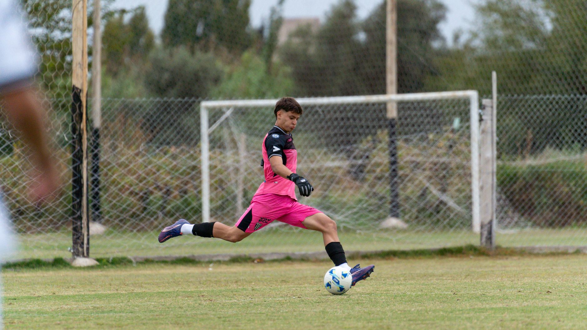 A young goalkeeper in pink uniform kicks a soccer ball on an outdoor field.