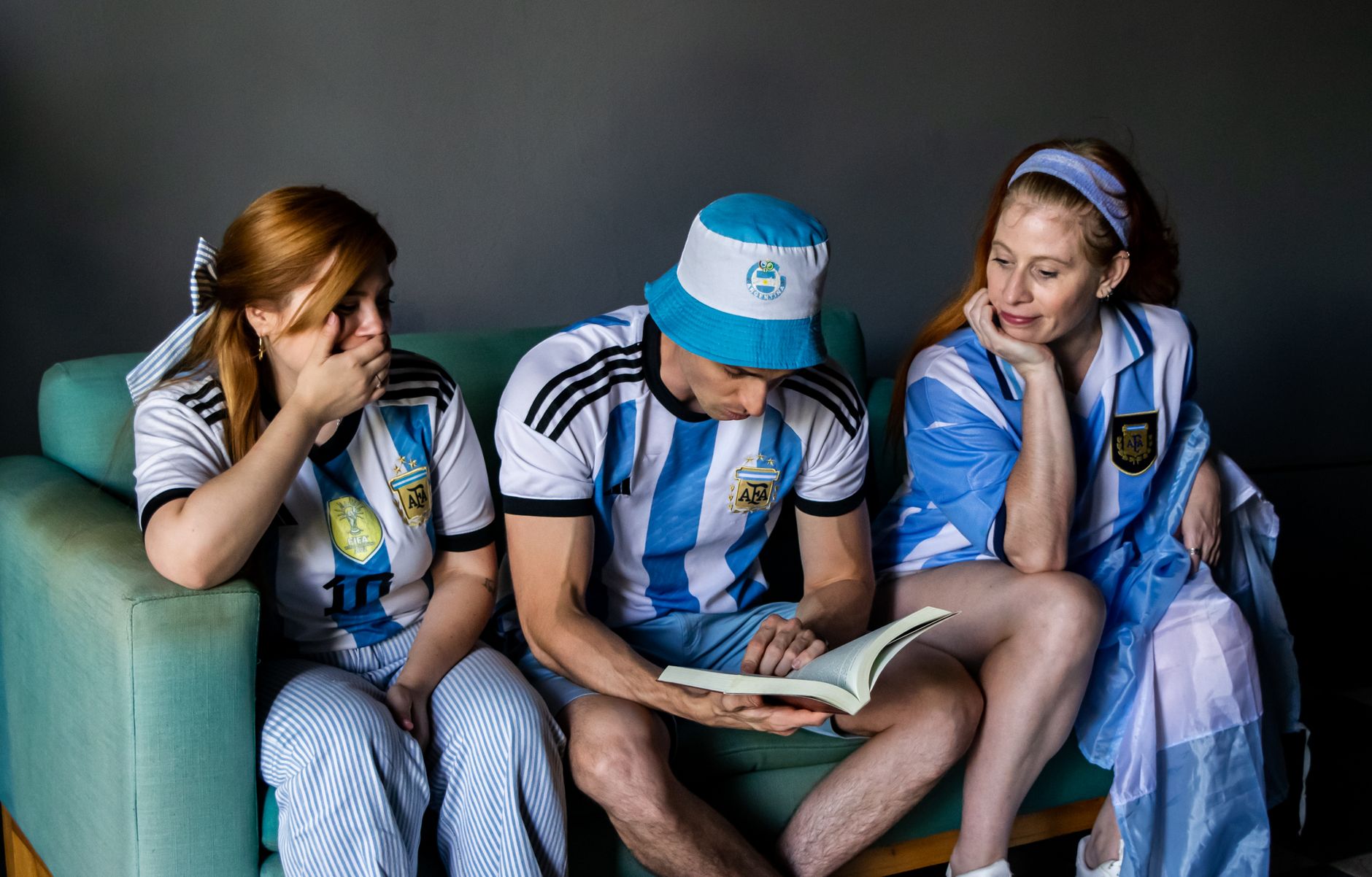 Three soccer fans in Argentina jerseys reading a book indoors