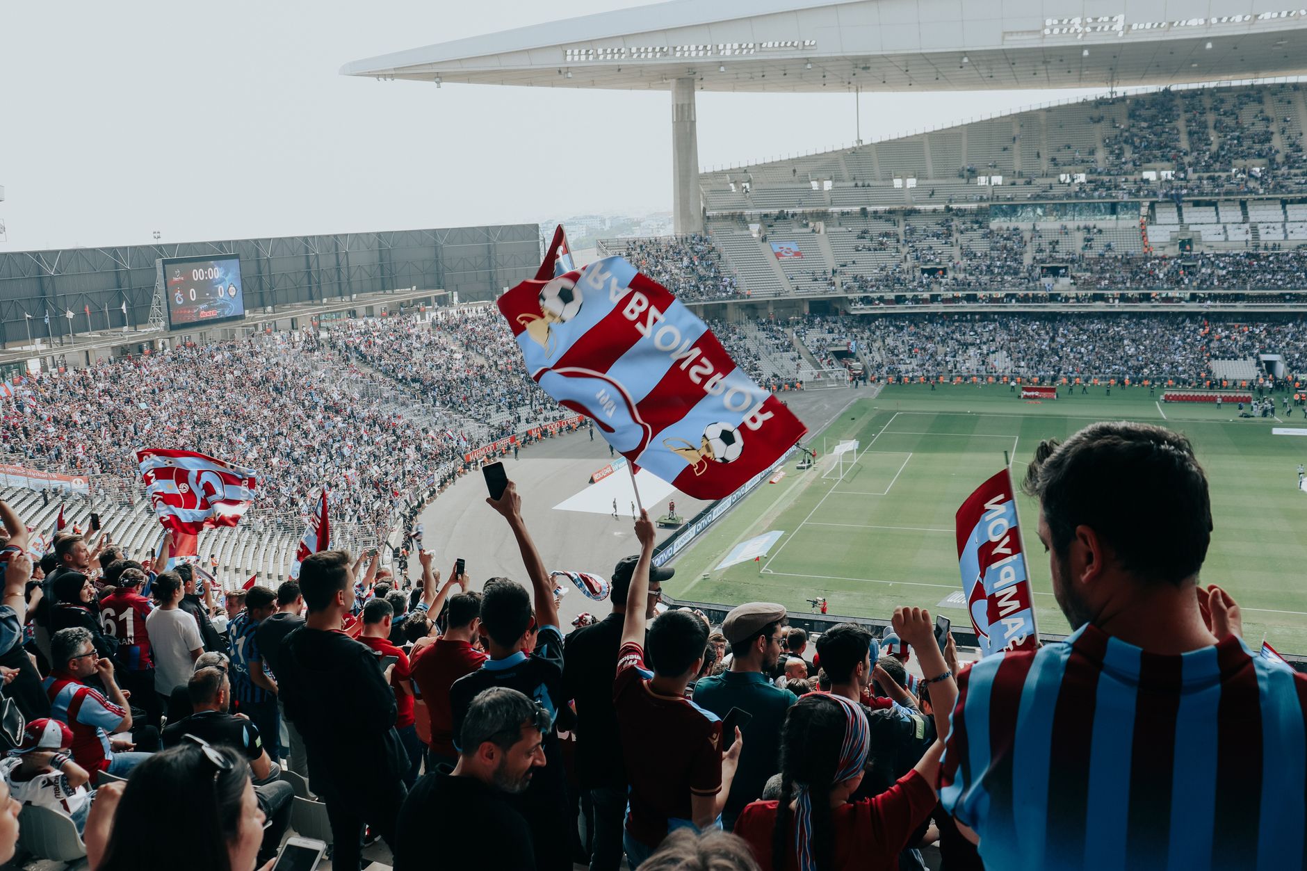 Vibrant crowd cheers at a football match in a large stadium, waving team flags.