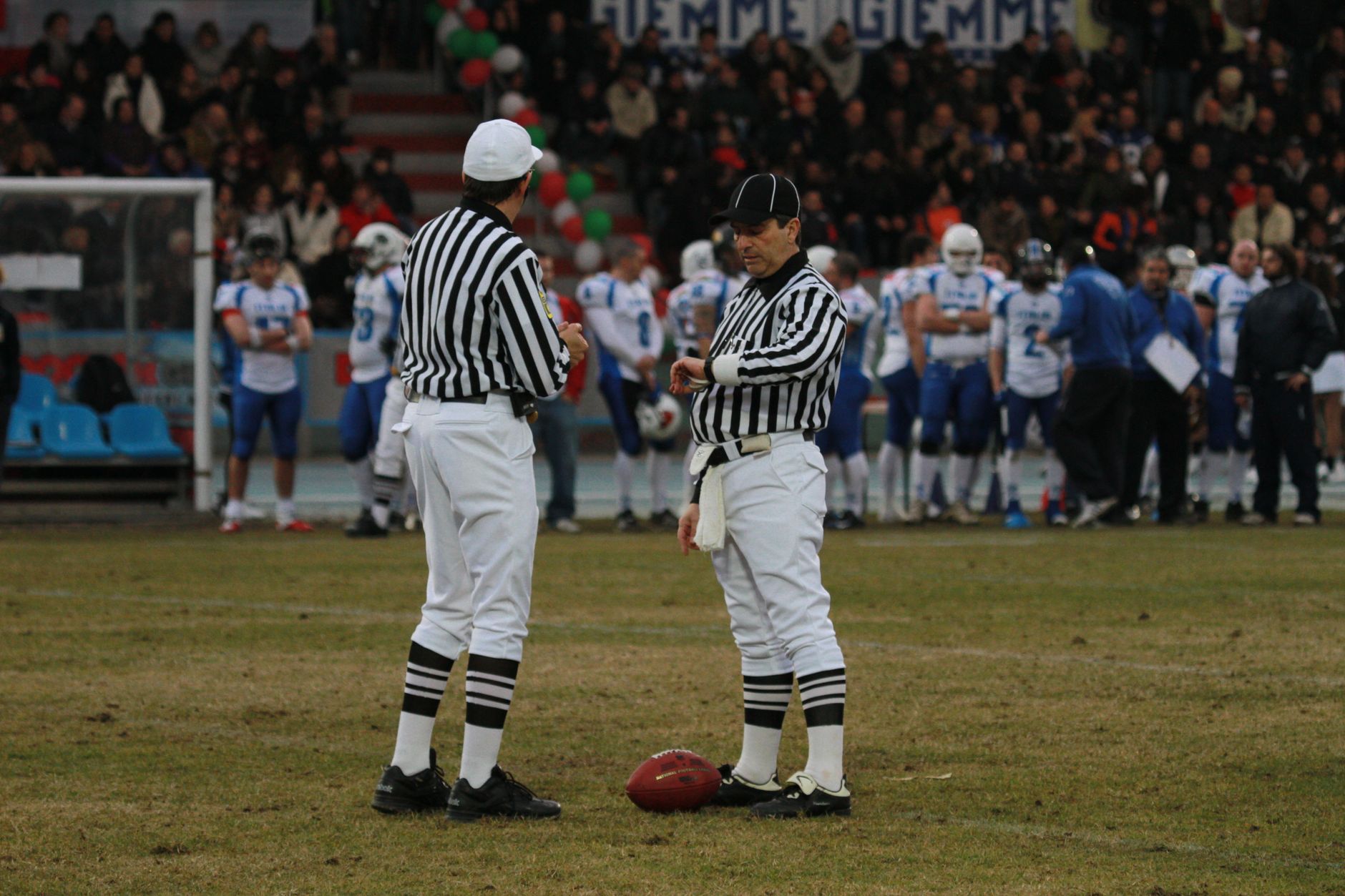 Two referees on a football field preparing for a game with players in the background at a stadium.