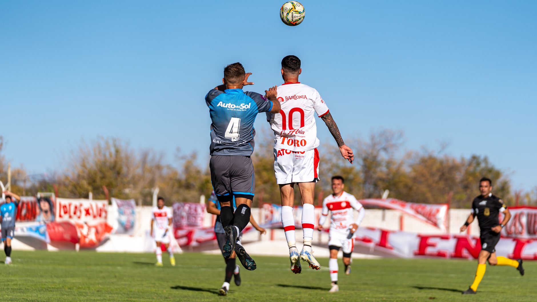 Soccer players in uniforms jumping to head the ball during an outdoor match on a sunny day.