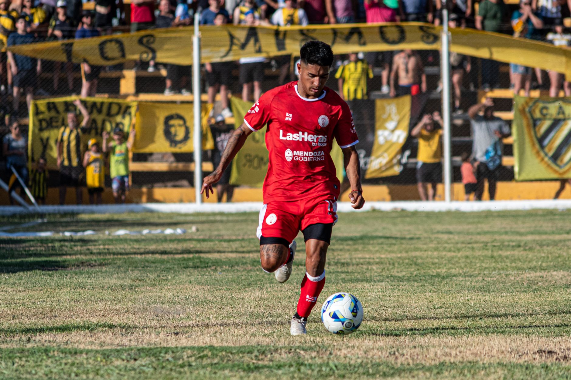 A soccer player in red uniform dribbling during a daytime match with spectators.