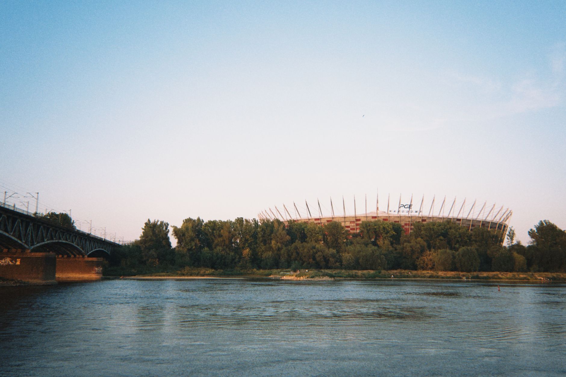 A serene view of the PGE Narodowy Stadium in Warsaw by the Vistula River during sunset.