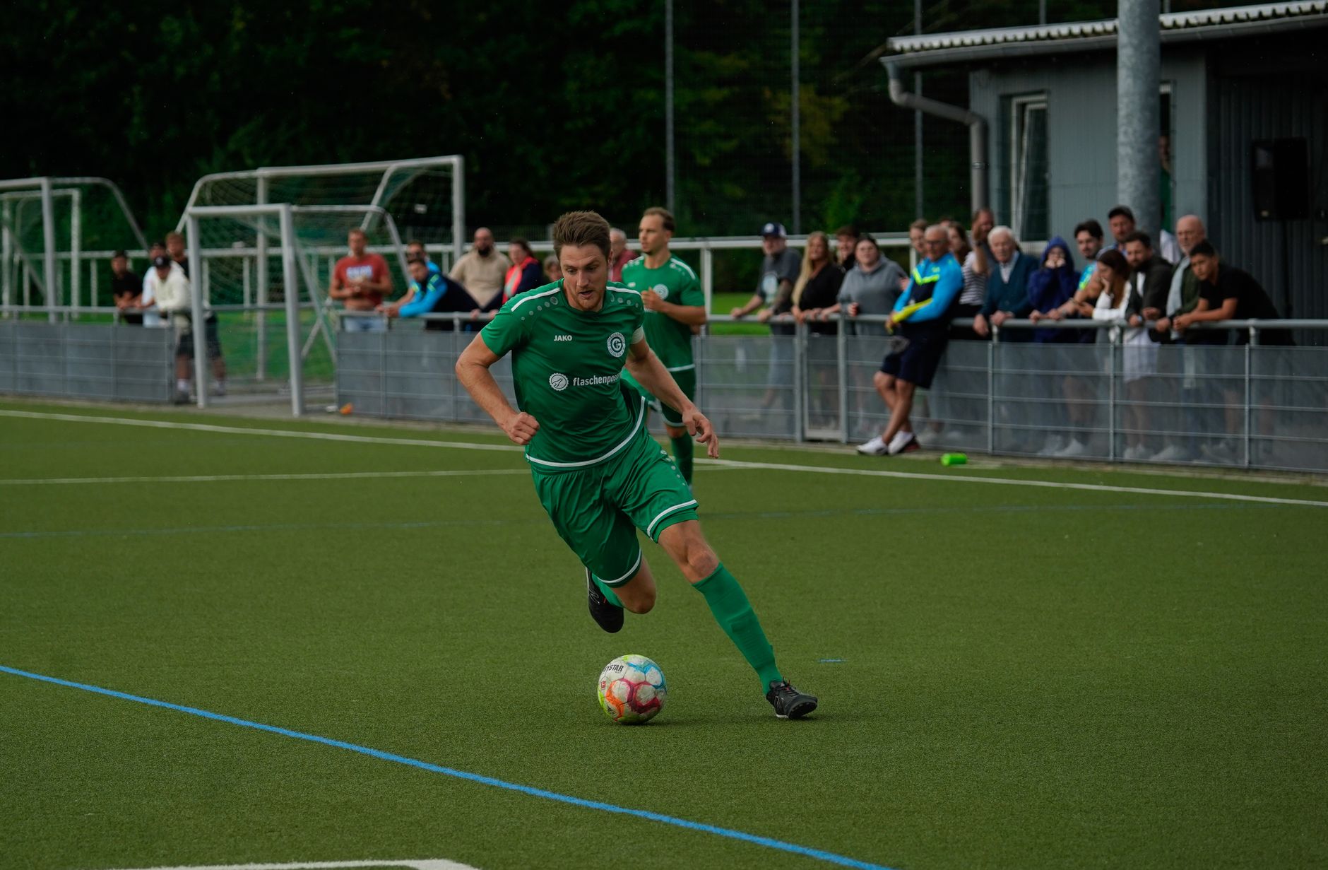 A soccer player in green dribbles the ball on a lively football pitch during a vibrant match.
