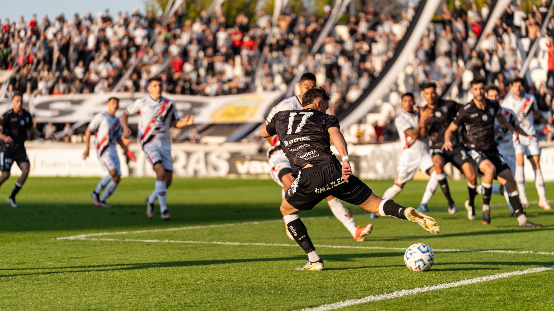 A dynamic football match with players in motion on a sunny day, witnessed by a packed stadium.