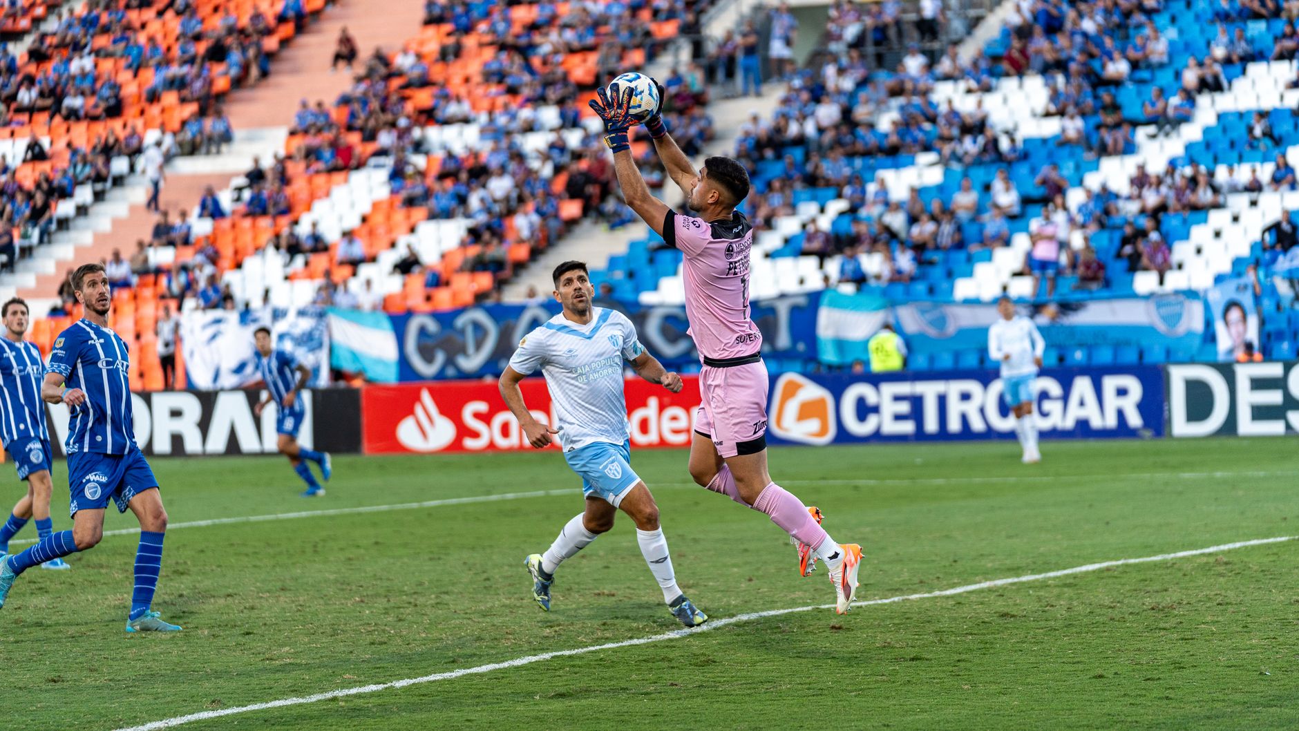 Goalkeeper in pink kit makes a save during a lively football match in a packed stadium.