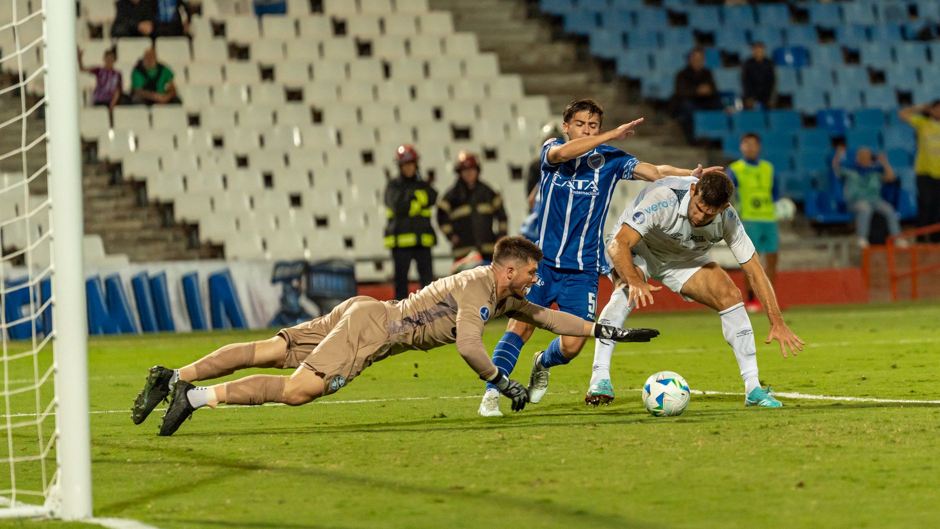 Dramatic soccer scene with goalkeeper diving to stop two players in a competitive match.